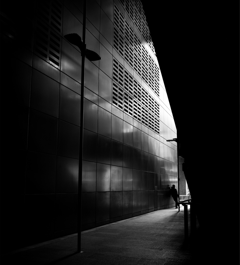Person walking along a dark sidewalk next to a reflective building facade at night.